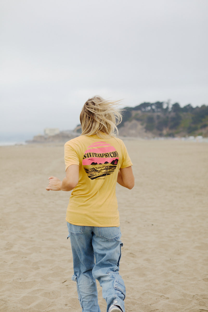 Person with long hair in a Kelly's Cove Tee walks on a sandy beach, facing away—capturing the true vibes of a coastal city.