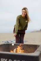 A woman in casual clothes and a cozy Elemental Crewneck smiles while roasting a marshmallow over a beach fire pit.