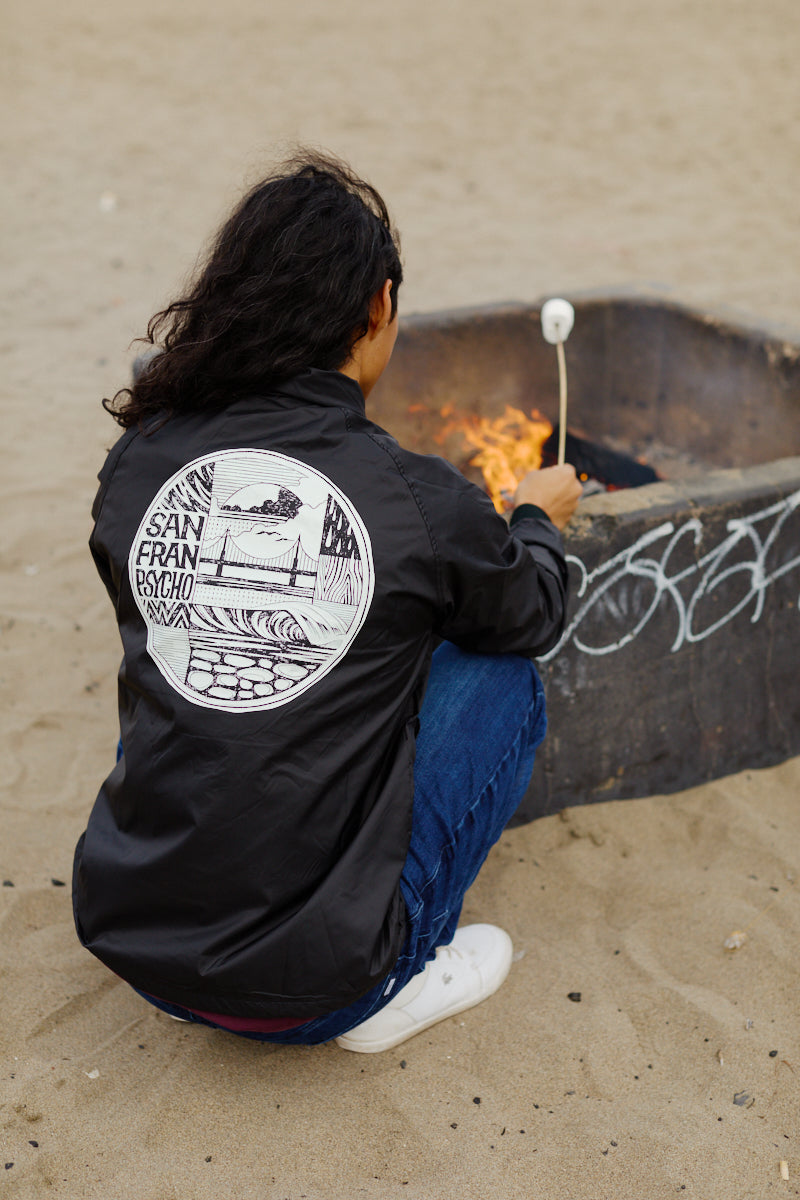 A person roasts a marshmallow over a beach fire pit, staying cozy in the wind-resistant Elemental Jacket featuring a San Francisco graphic.