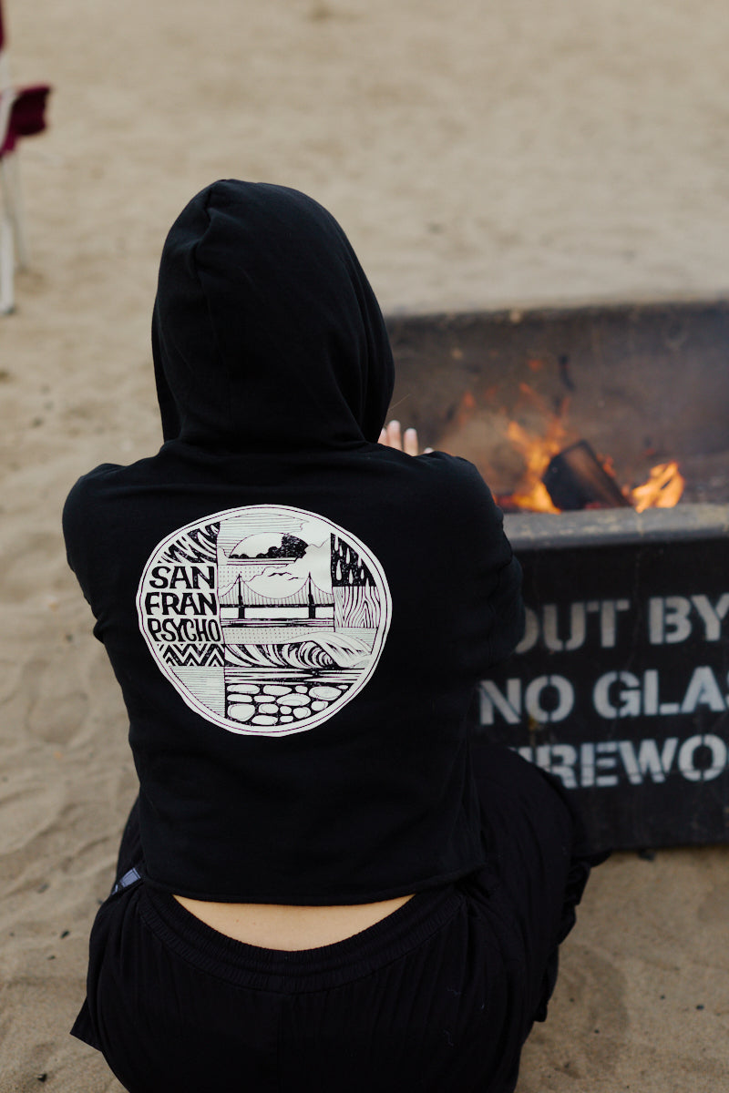 Someone in an Elemental Crop Hoodie featuring a circular San Franpsycho design sits by a beach fire pit.