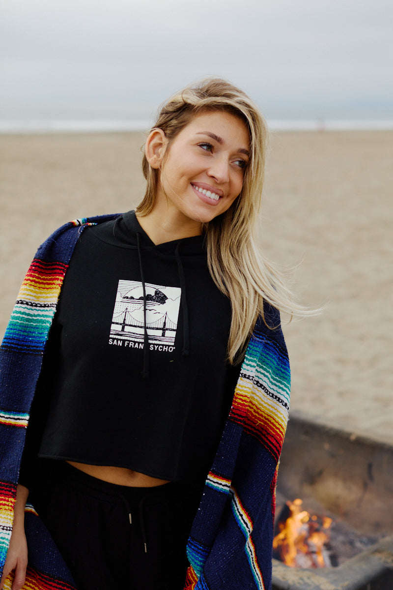 A smiling woman wearing the Elemental Crop Hoodie and holding a colorful blanket stands by a fire pit on a sandy beach.
