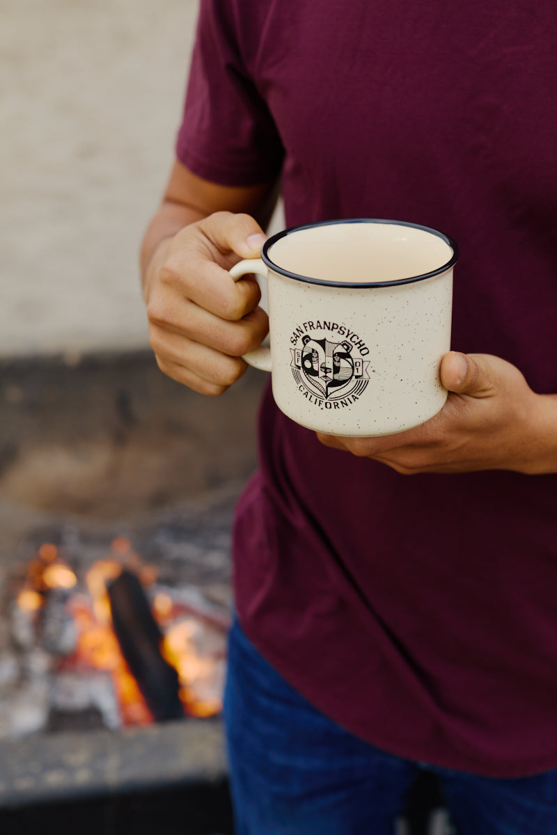 A person in a maroon shirt holds the Bear Essentials Mug—a speckled ceramic mug featuring San Francisco, California—by a campfire, capturing a cozy West Coast vibe.