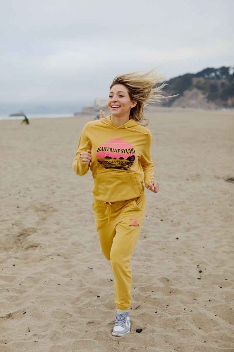 A woman jogs along the sandy beach, smiling with windblown hair and easy coastal vibes in a hand-printed Kelly's Cove Pullover Hoodie.