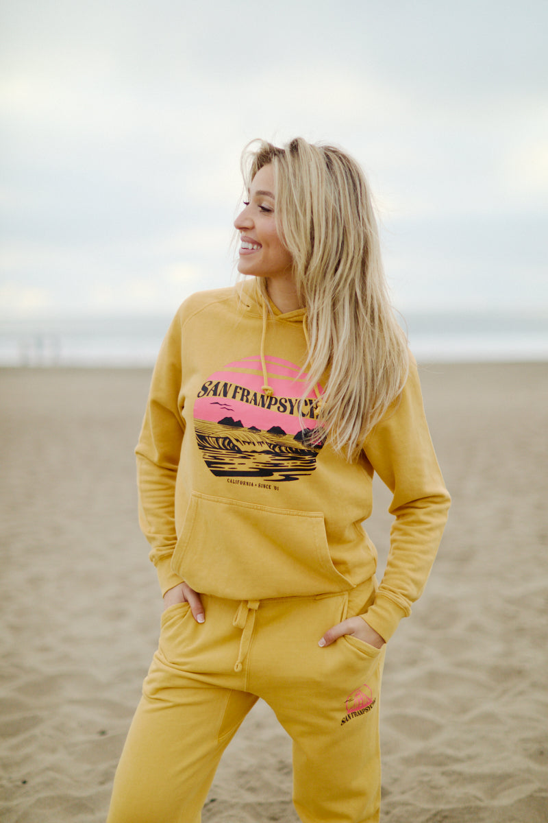 A woman in a hand-printed yellow Kelly's Cove Pullover Hoodie smiles while standing on a sandy beach, capturing coastal city vibes.