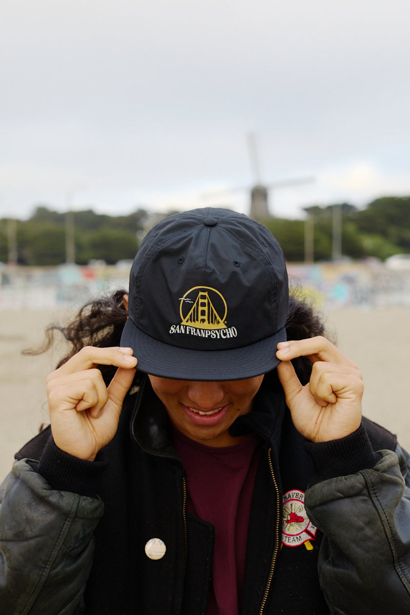 A person smiles and holds the flat brim of a Gull Logo Hat with an unstructured crown, as a windmill stands in the background.