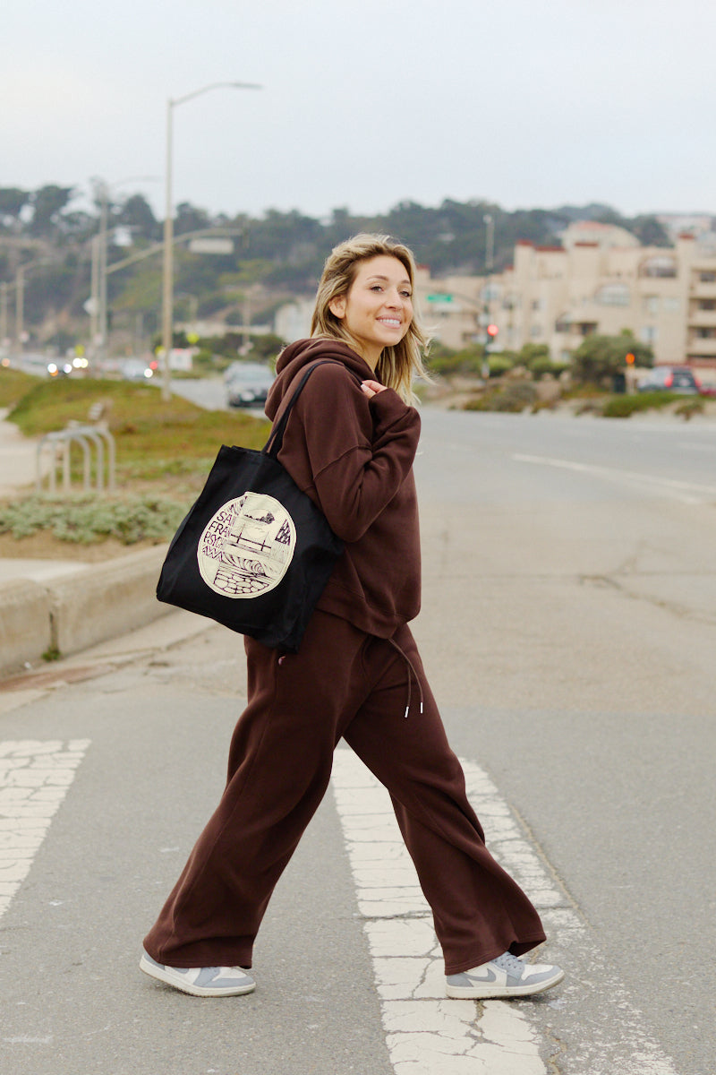 A woman in a brown tracksuit smiles while crossing a street, carrying the Elemental Tote featuring bold Erik Abel artwork.