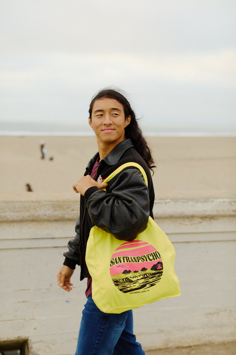 A person with long hair smiles at the camera on a San Francisco beach, holding a yellow Kelly's Cove Tote by San Franpsycho.