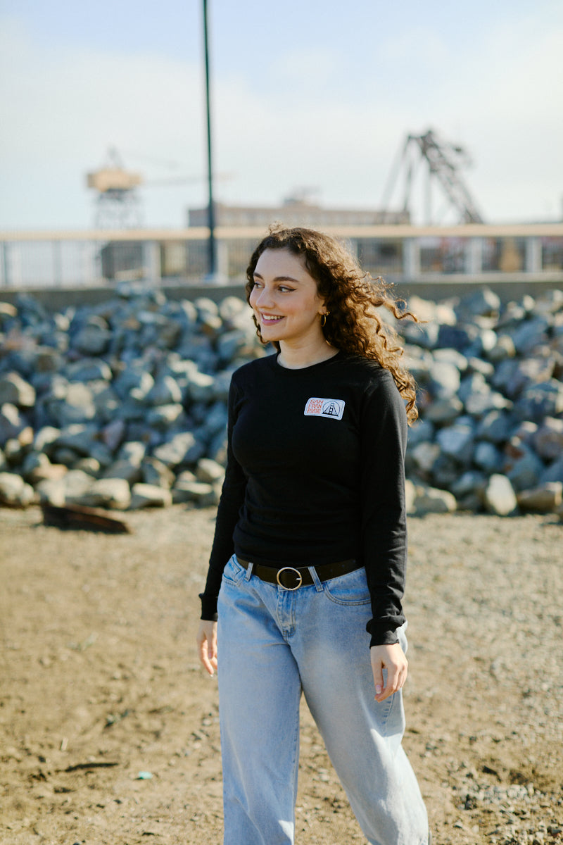 Smiling woman with curly hair in a black Elemental Long Sleeve and jeans stands outdoors near rocks on a sunny day.