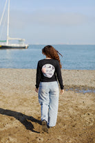 A woman with curly hair walks on a sandy beach toward the sea, dressed in jeans and the Elemental Long Sleeve.