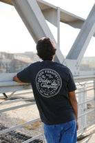 Wearing the Salt of the City Tee, hand printed in San Francisco, a person in jeans leans on a bridge railing, facing away from the camera.