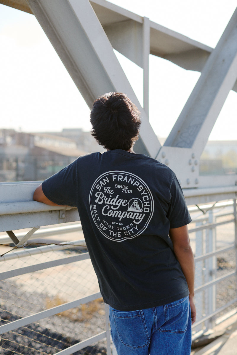 Wearing the Salt of the City Tee, hand printed in San Francisco, a person in jeans leans on a bridge railing, facing away from the camera.
