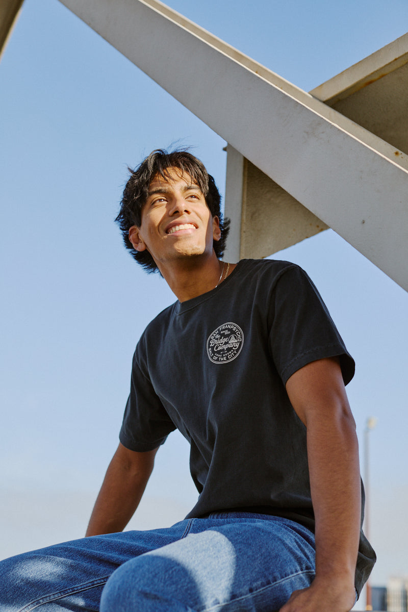 A young man in the Salt of the City Tee and jeans sits outdoors, smiling as he looks up under a metal structure.
