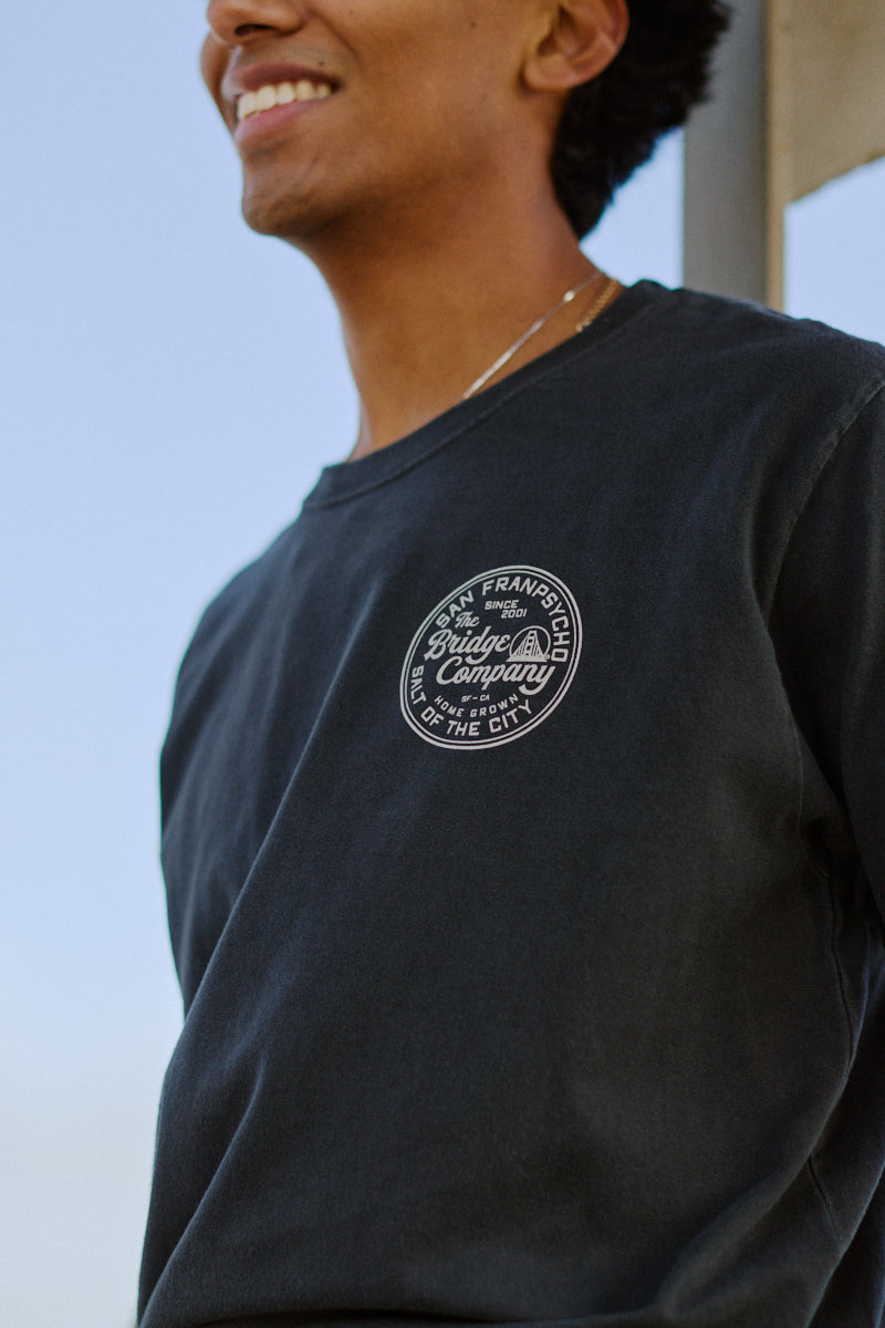Smiling person wearing the Salt of the City Tee, a dark heavyweight cotton shirt with a round Straight Up Company logo hand-printed in San Francisco on the chest.