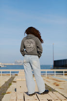 A person with curly hair in jeans and a Salt of the City Pullover Hoodie stands on dockside planks, gazing at the ocean under a clear sky.