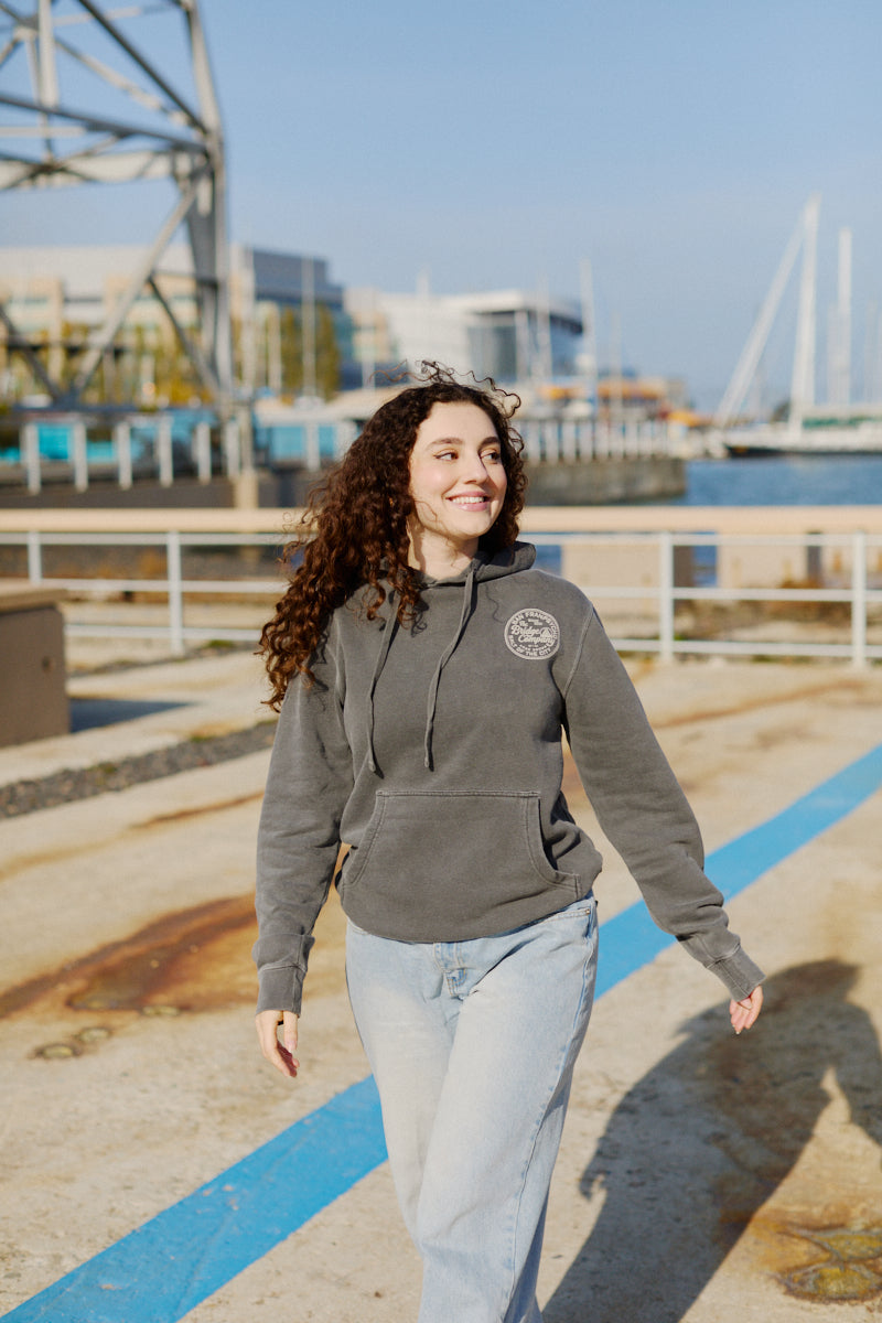 A smiling woman wears the Salt of the City Pullover Hoodie while walking outdoors near a waterfront on a sunny day.