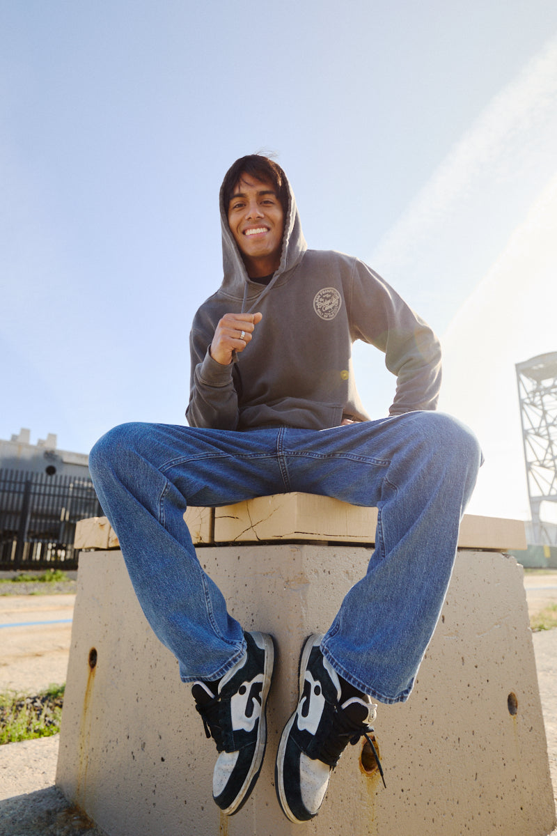 A smiling young man wearing the Salt of the City Pullover Hoodie and jeans sits on a concrete block outdoors, sunlight shining behind him.