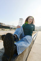 A woman with curly hair sits on a concrete ledge outdoors, smiling in her Bridge Co Tee, one booted foot pointed toward the camera.