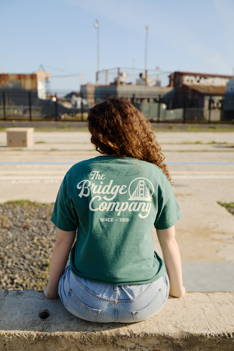 A person with curly hair sits outdoors, wearing a green hand-printed Bridge Co Tee and denim shorts.