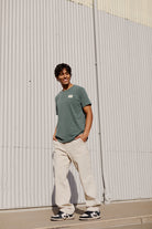 A person wearing the Bridge Co Tee in green stands outside against a corrugated metal wall, smiling.