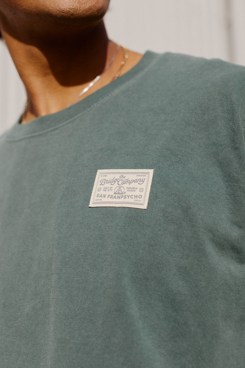 A person wears the Bridge Co Tee, a heavyweight green cotton shirt with a beige "The Bridge Company, San Franpsycho" patch label on the chest—ideal for workwear style.
