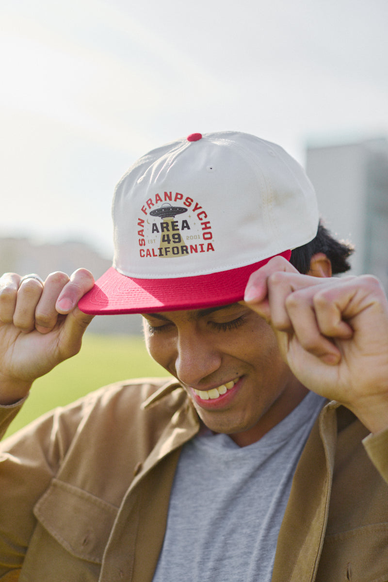 Smiling person adjusts Area 49 Hat, featuring a white and red design with the Area 49 Logo and adjustable back, outdoors in California.