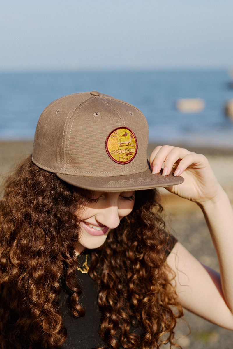 A smiling woman with curly hair wears the Elemental Hat by the beach, adjusting its flat brim with her hand.