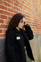 A young woman with curly hair in the Bridge Co. Chore Jacket leans against a brick wall, looking down thoughtfully—embodying the timeless spirit of San Francisco-made workwear.