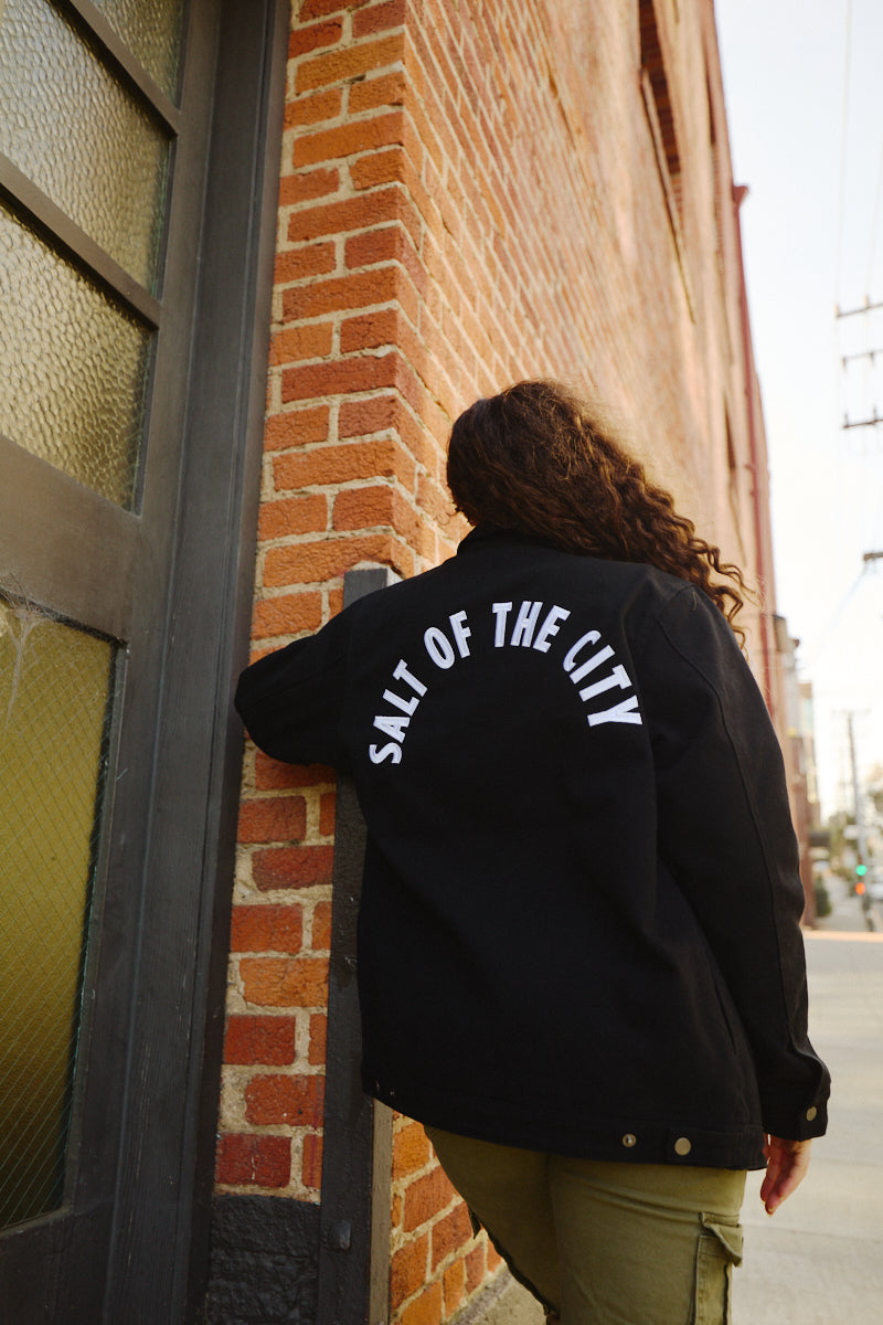 A person with curly hair stands by a brick wall wearing the Bridge Co. Chore Jacket in black, featuring "SALT OF THE CITY" text and made in San Francisco.