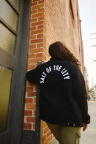 A person with curly hair stands by a brick wall wearing the Bridge Co. Chore Jacket in black, featuring "SALT OF THE CITY" text and made in San Francisco.