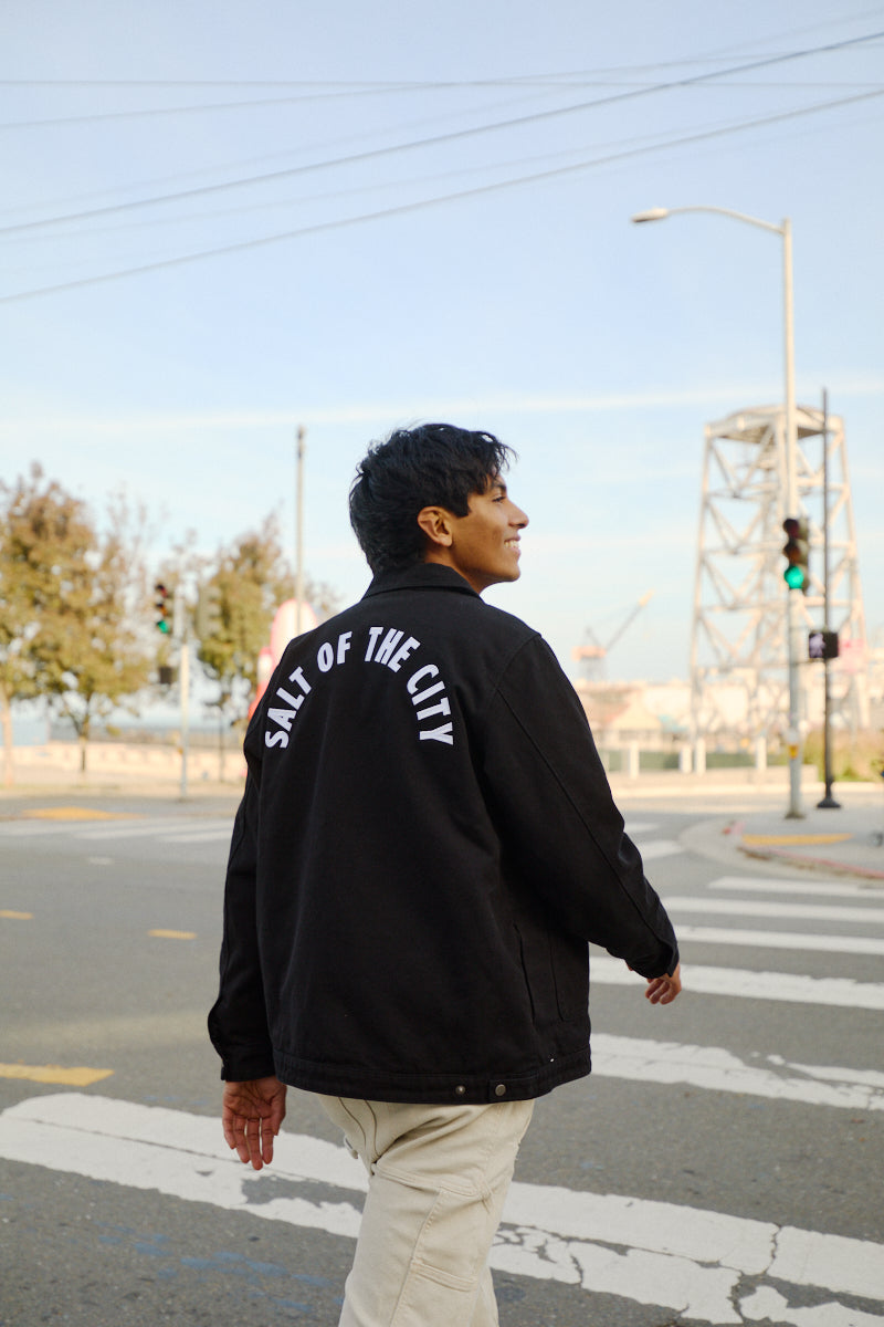 A person crosses the street in a black Bridge Co. Chore Jacket with SALT OF THE CITY on the back, capturing classic workwear style made in San Francisco.