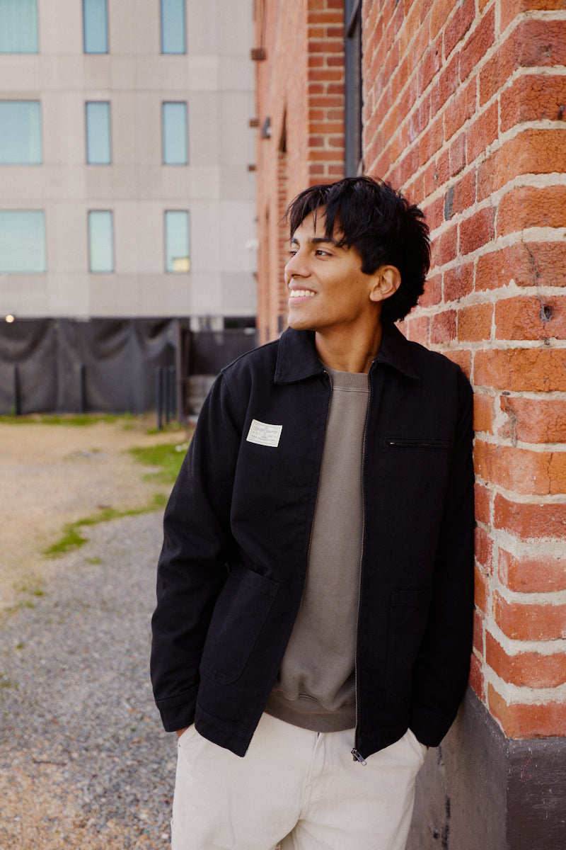 A young man in a Bridge Co. Chore Jacket smiles as he leans against a red brick wall outdoors, highlighting classic workwear style.