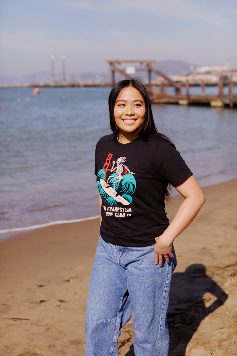 A woman smiles on a sandy beach wearing jeans and the Surf Club Tee, featuring a comfy design with a black San Francisco Surf Club logo.