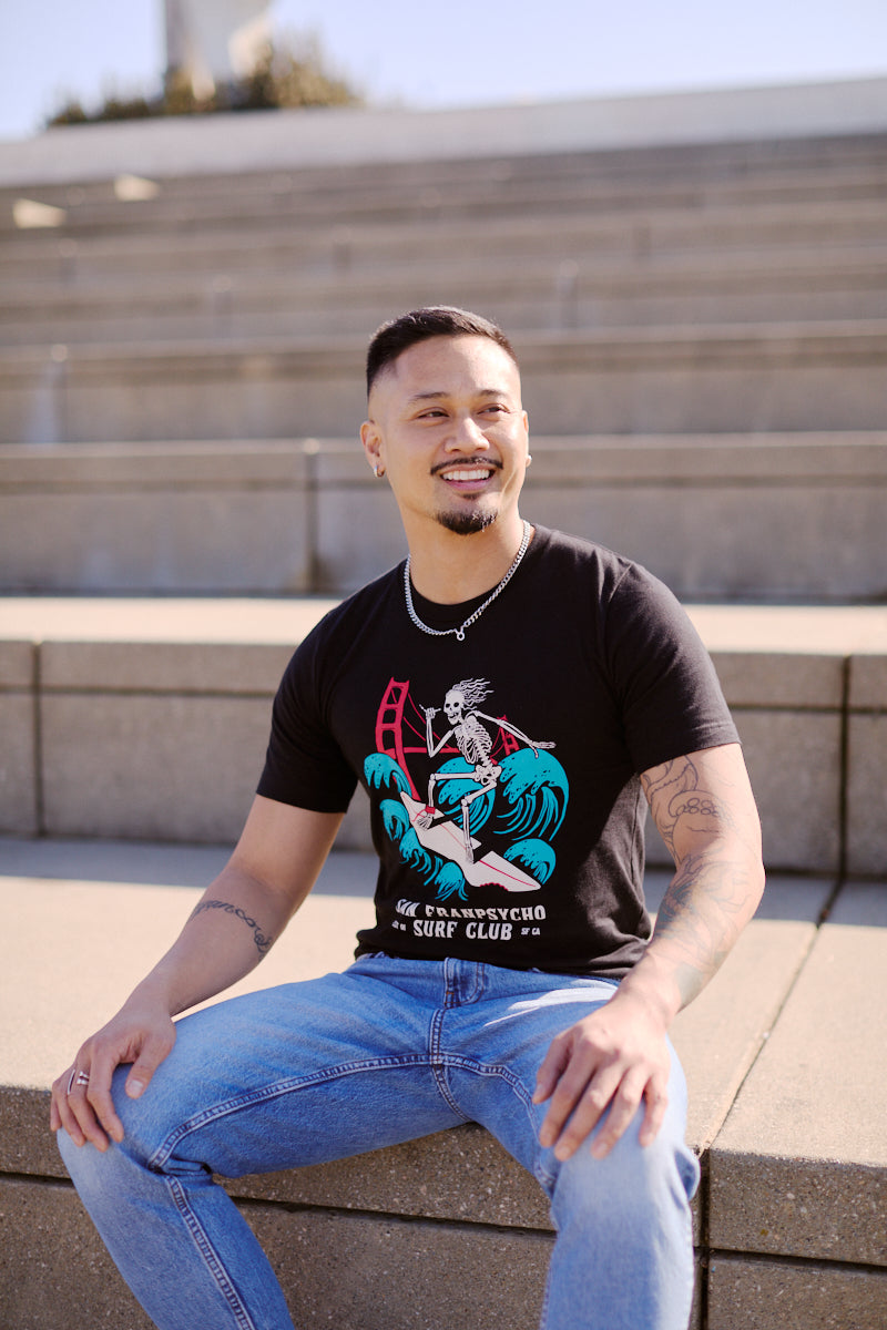 A man wearing a Surf Club Tee and jeans sits on outdoor steps, smiling and looking to the side.