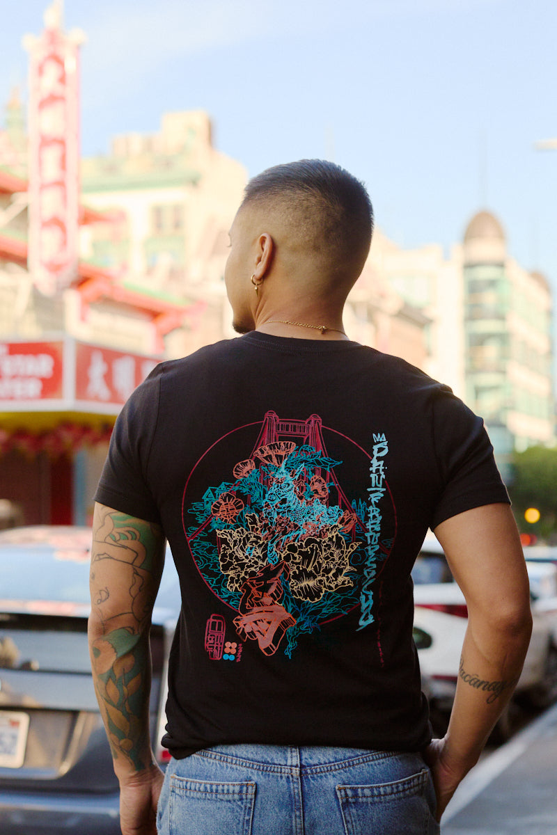 A tattooed man stands on a city street with colorful buildings, wearing the Year of the Horse Tee—a black graphic t-shirt made from soft Airlume cotton.