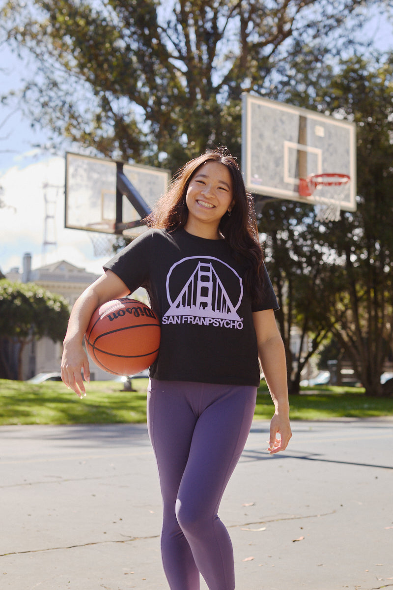 Smiling woman holding a basketball on an outdoor court, wearing the Women's Black & Purple Logo Crop Tee and purple leggings.