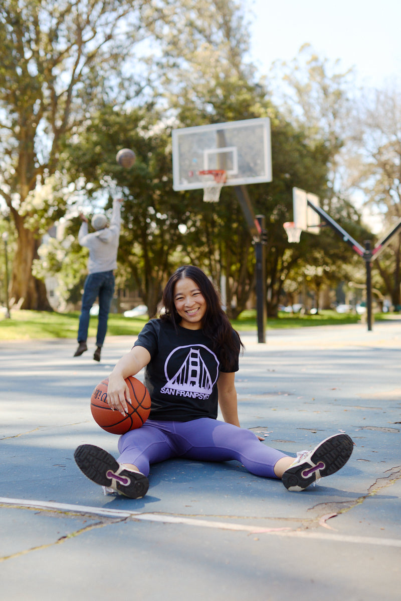 A young woman in a Women's Black & Purple Logo Crop Tee sits on an outdoor basketball court, smiling with a basketball, as someone shoots a basket behind her.