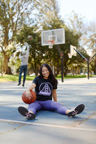 A young woman in a Women's Black & Purple Logo Crop Tee sits on an outdoor basketball court, smiling with a basketball, as someone shoots a basket behind her.