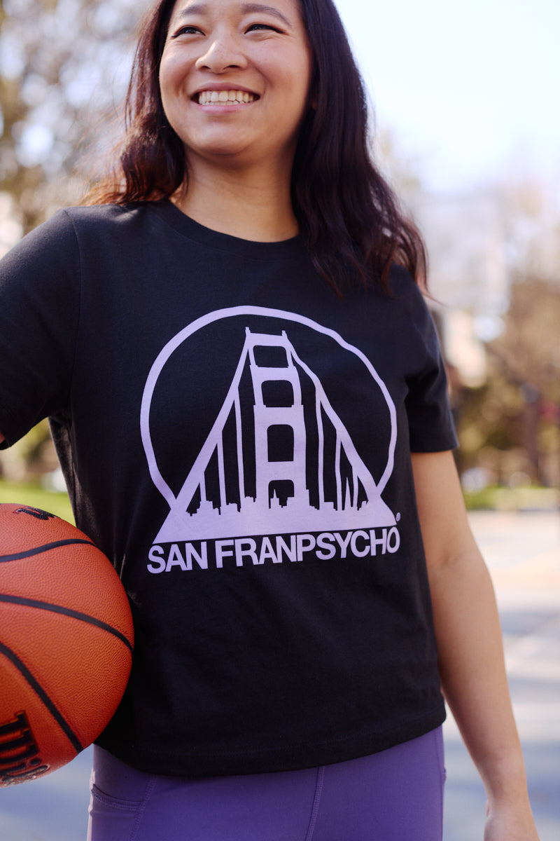 Smiling woman outdoors holding a basketball, wearing a Women's Black & Purple Logo Crop Tee.