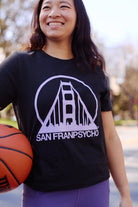 Smiling woman outdoors holding a basketball, wearing a Women's Black & Purple Logo Crop Tee.