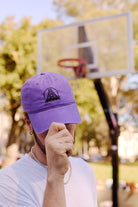 A person wearing the Purple & Black Logo Dad Hat stands by an outdoor basketball hoop, holding the brim of the comfortable hat down.