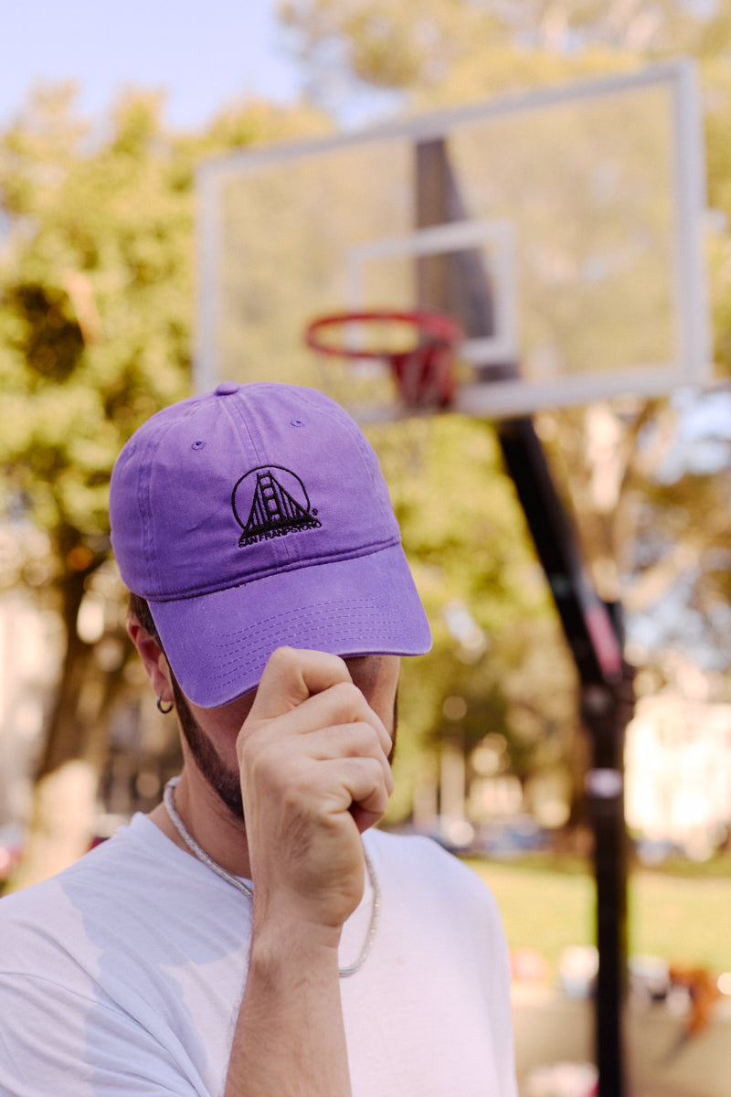 A person wearing the Purple & Black Logo Dad Hat stands by an outdoor basketball hoop, holding the brim of the comfortable hat down.