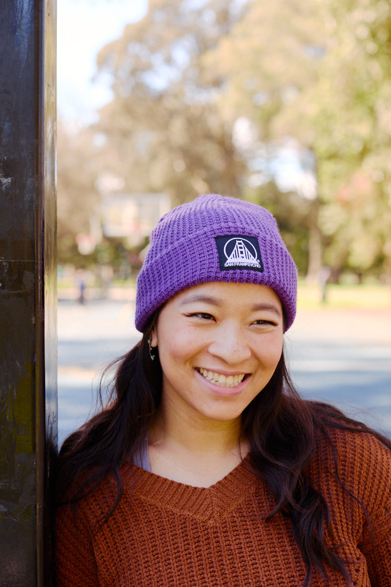A woman wearing the Purple Waffle Beanie w/ Black/White Logo and a brown sweater leans against a pole outdoors on a sunny day.