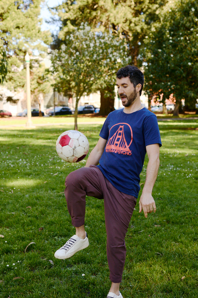 A man in a Navy & Poppy Logo Tee balances a soccer ball on his knee in a sunny park with trees in the background.