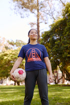 Person wearing a Navy & Poppy Logo Tee holds a soccer ball while standing on grass in a sunny park.