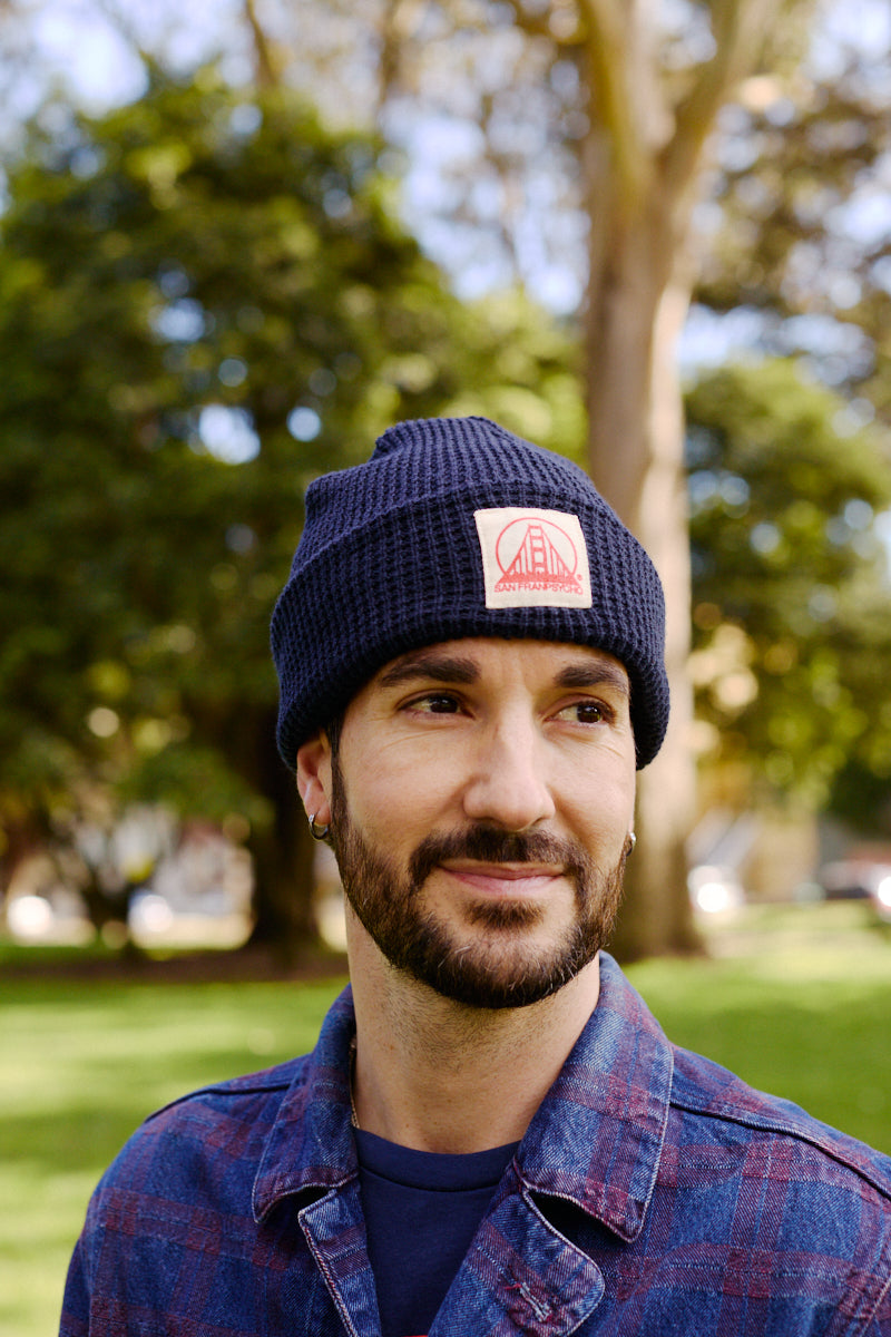 A bearded man in a plaid shirt stands outside in a sunny, green park, wearing a Navy Waffle Beanie with Natural/Poppy Logo.