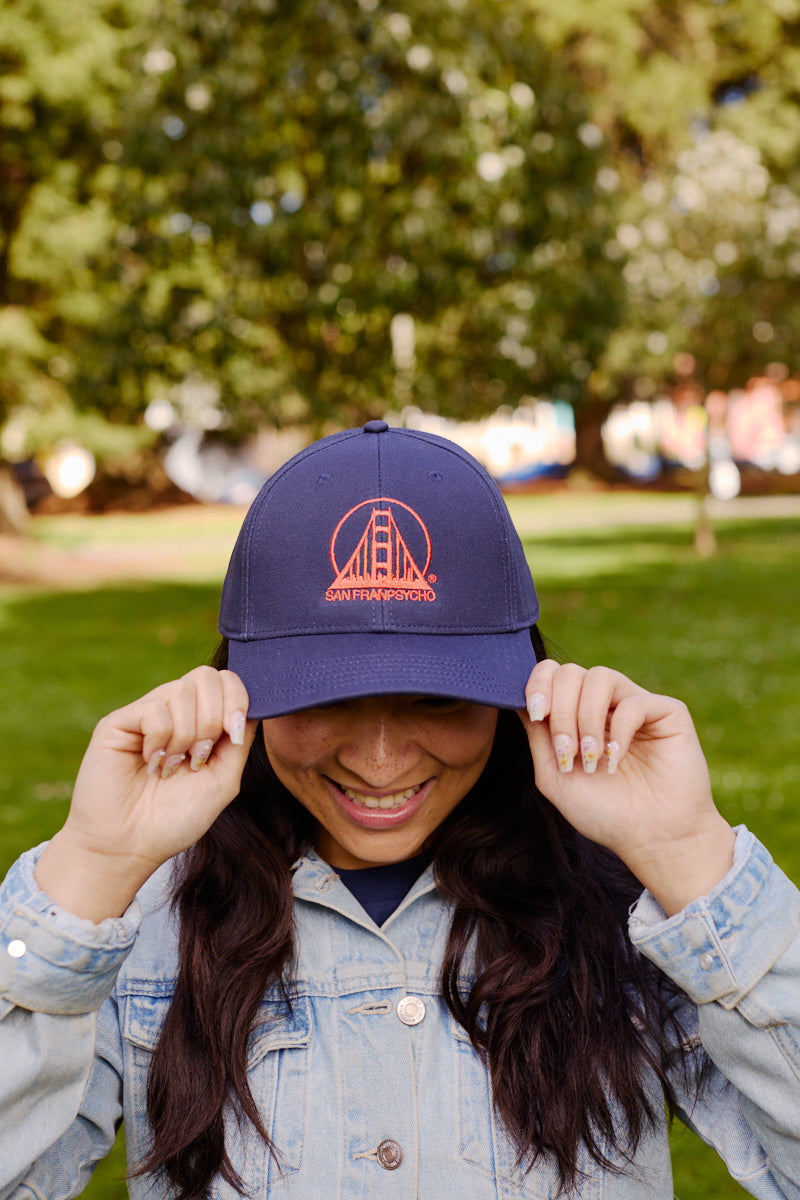 A smiling person in a denim jacket holds the Embroidered Logo Snapback - Navy & Poppy outdoors in a sunny, green park.