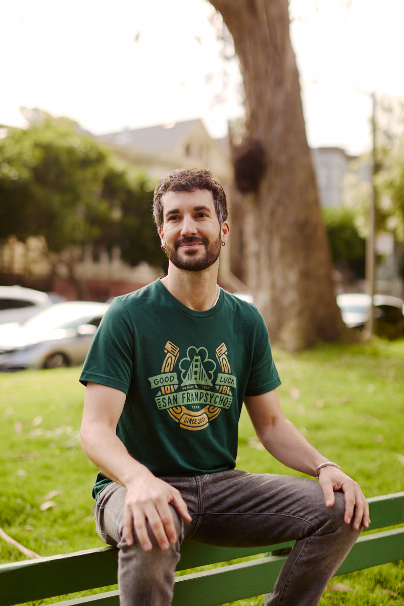Man with short dark hair and a beard sits on a green park bench, wearing the Lucky Psycho Tee, a hand-printed St. Paddy’s shirt featuring San Francisco.