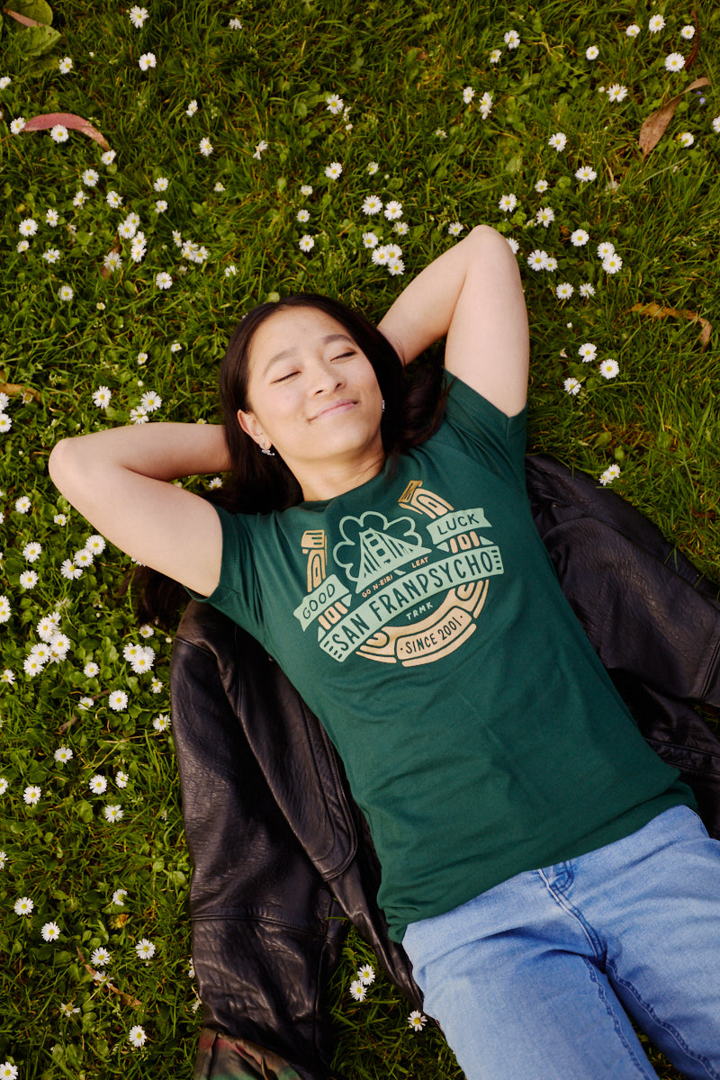 Woman lies on grass among white flowers, smiling with eyes closed in a hand-printed Lucky Psycho Tee and blue jeans—celebrating St. Patrick’s Day in style.