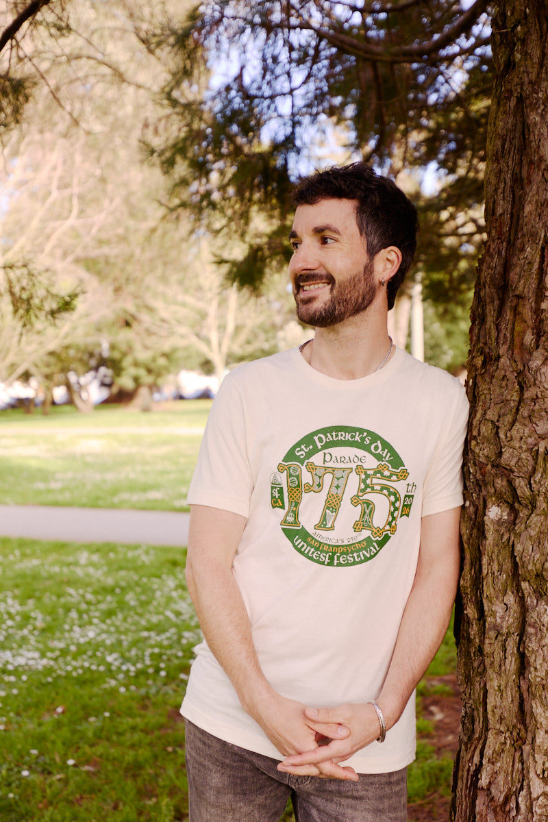 Man smiling and leaning against a tree in a park, wearing the 175th St Patrick's Day Parade Tee, a white limited edition commemorative shirt designed for the festival.