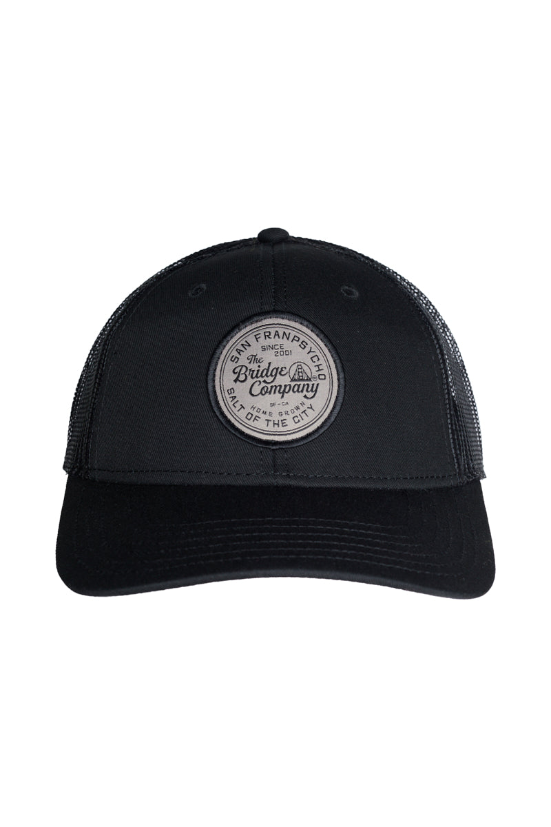 The Salt of The City Trucker Hat is a black cap with a round patch on the front reading The Bridge Company, San Francisco. It features an adjustable back for comfort and fits most sizes.